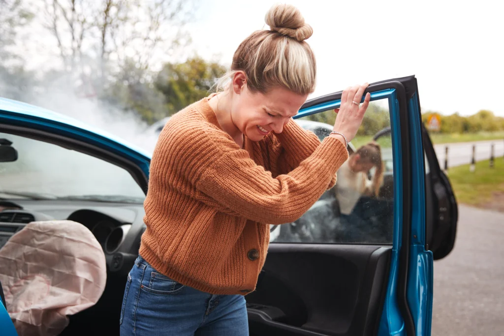 Woman grimacing in pain while leaning on a damaged car door with a deployed airbag and smoke rising from the hood, following a car accident.