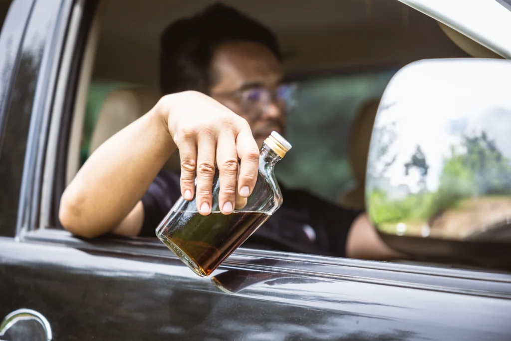 Person extending a liquor bottle out of a moving car window with blurred greenery reflected in the side mirror, symbolizing dangerous drunk driving behavior.