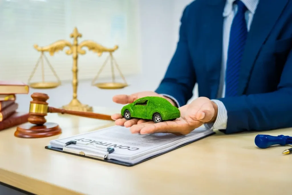 A lawyer in a blue suit and tie holds a small, green, moss-covered toy car in his open hands, positioned above a clipboard with a "CONTRACT" document. On the wooden desk, a golden scale of justice, a gavel, and books are visible, along with a pen and a blue stamp. The image conveys themes of environmental law, green initiatives, car-related legal matters, or sustainable business.