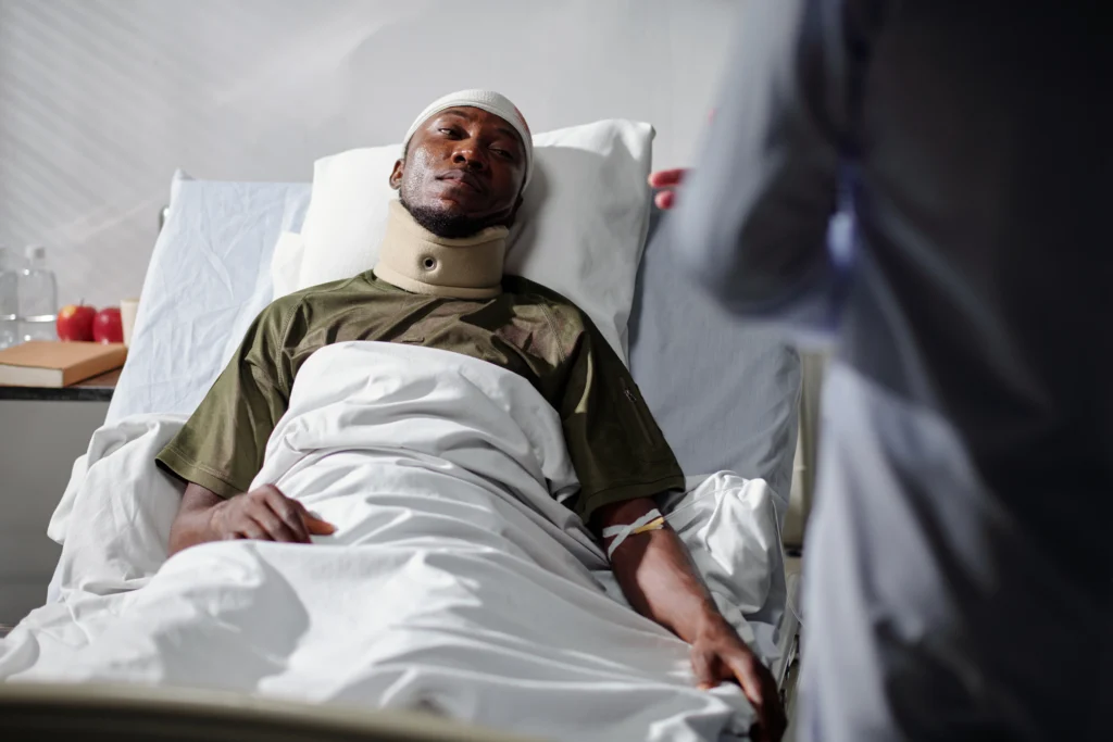 Man with a bandaged head and neck brace lying in a hospital bed, connected to an IV, with a visitor standing nearby, symbolizing recovery after a motorcycle accident.