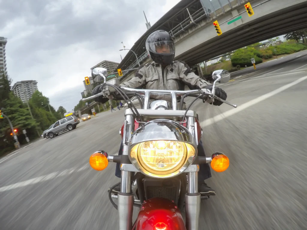 Motorcyclist wearing a gray jacket and helmet riding under an overpass on a city street with traffic lights, surrounded by urban buildings and an overcast sky, emphasizing the risks motorcyclists face in urban environments.