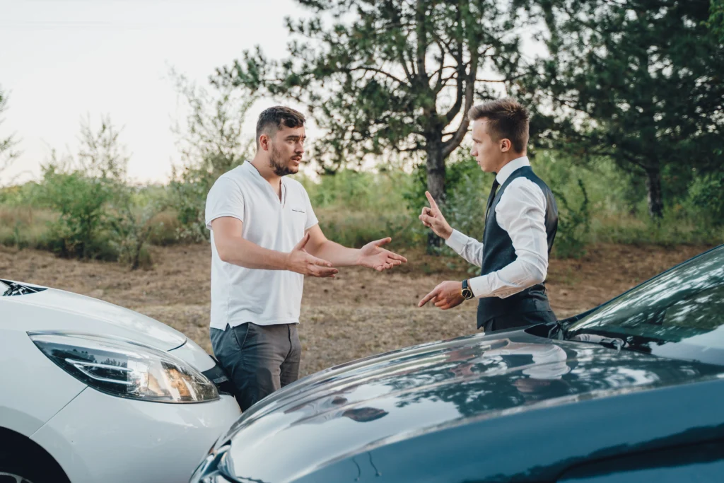 Two men arguing in front of damaged vehicles on a rural road, one in a white shirt gesturing emphatically and the other wearing a vest, illustrating a dispute over accident responsibility.