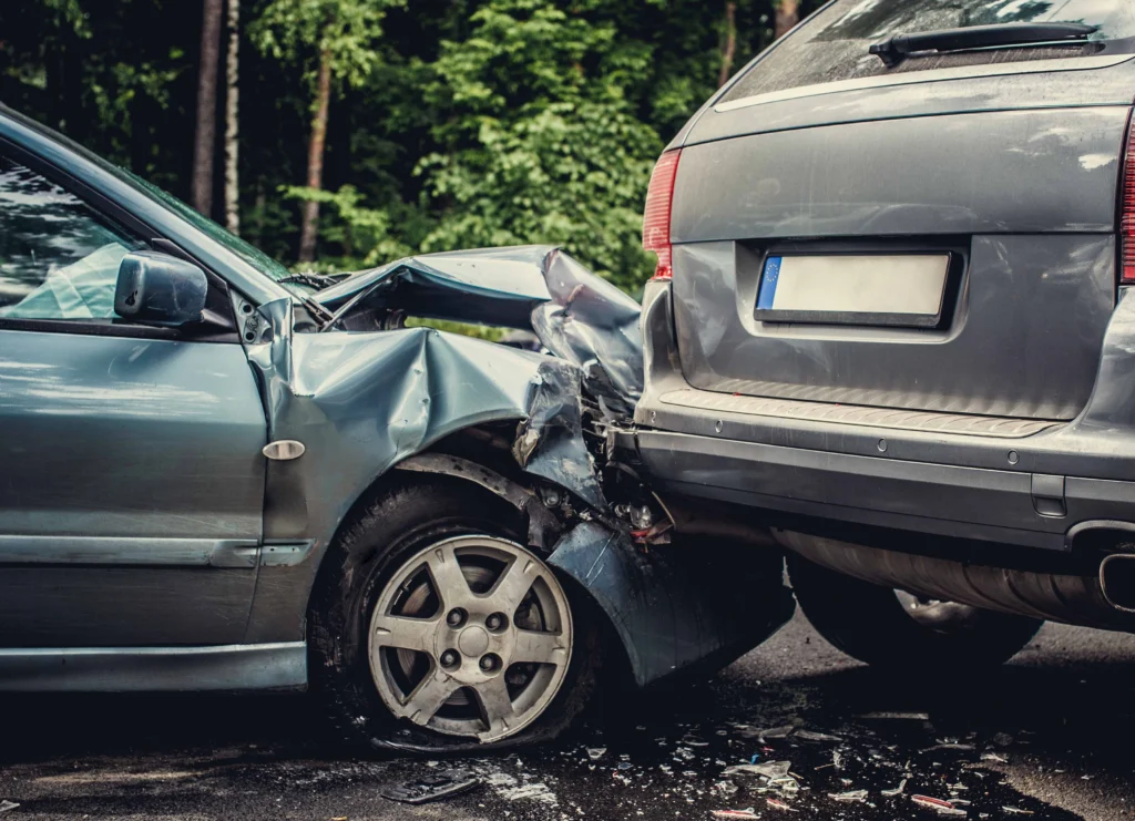 Two cars in a rear-end collision on a forest road, blue car’s front damaged against gray car’s rear with debris scattered.