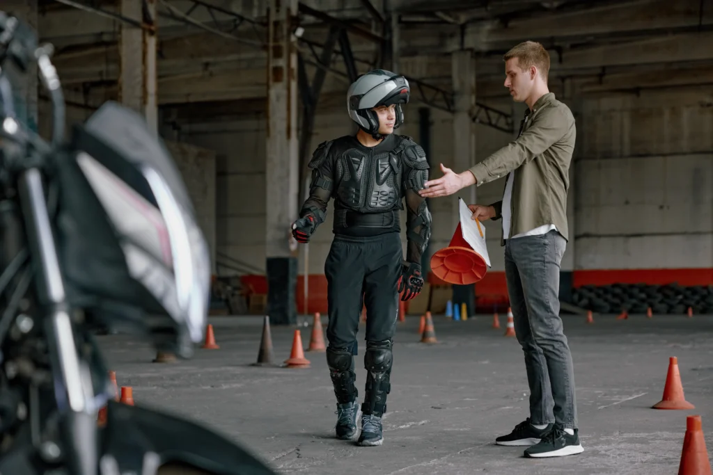 A person in motorcycle gear listens to an instructor holding cones and papers during a motorcycle safety training session.