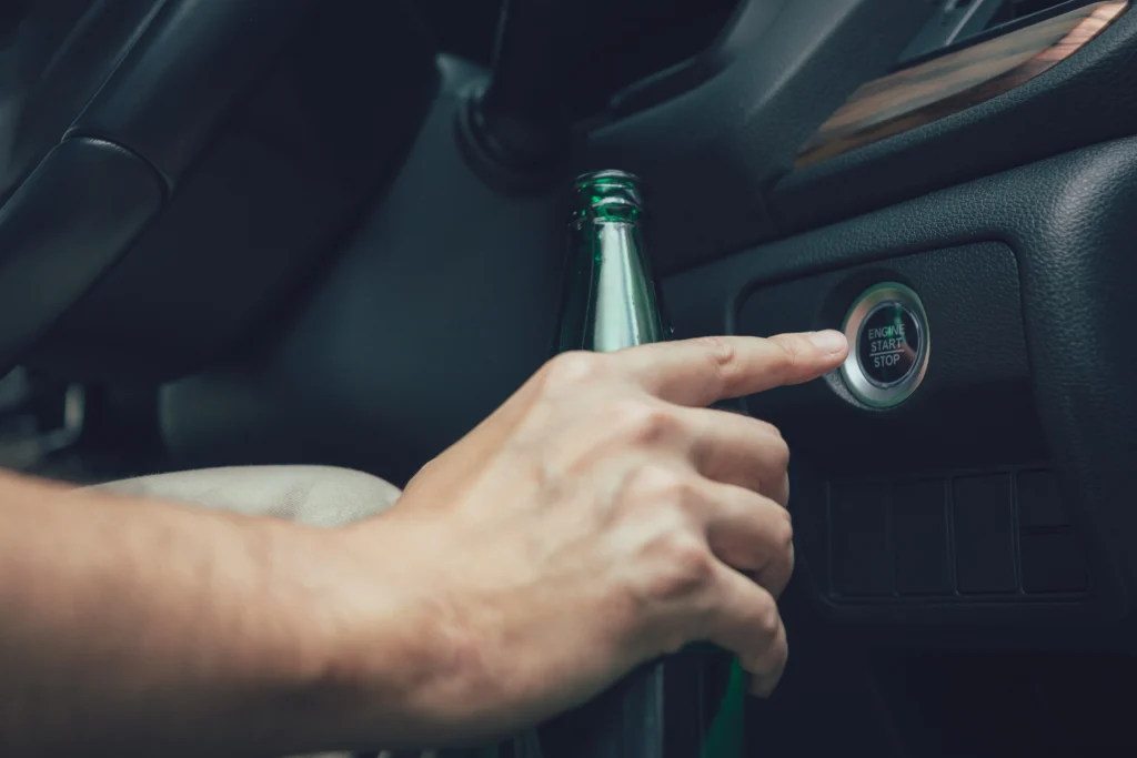 A hand holding a liquor bottle presses a car's "Engine Start Stop" button on the dashboard. The tone is casual and the setting is a vehicle interior.