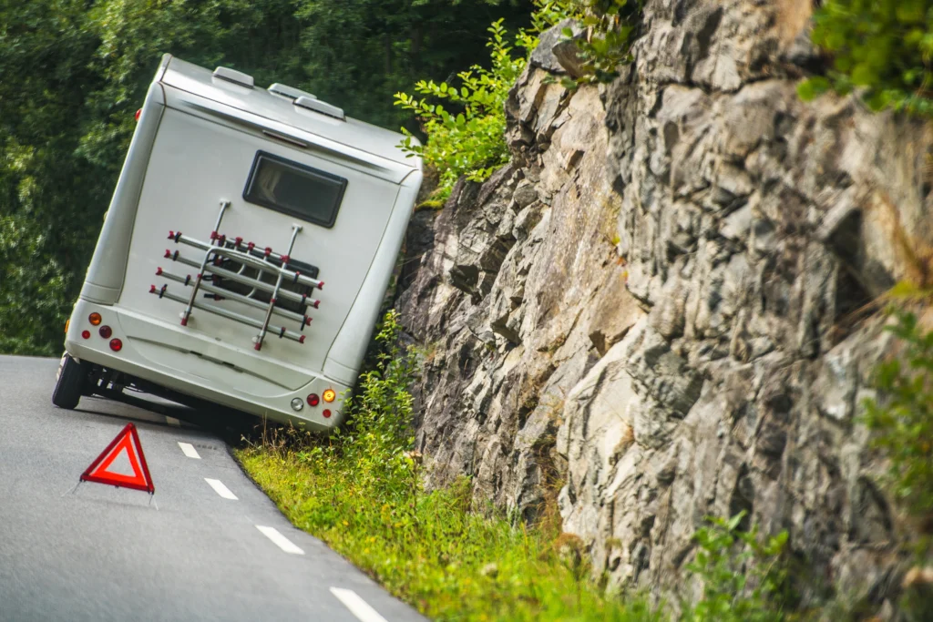 Camper van leaning against a rocky hillside on a narrow road, with a red warning triangle placed behind it, suggesting a roadside emergency.