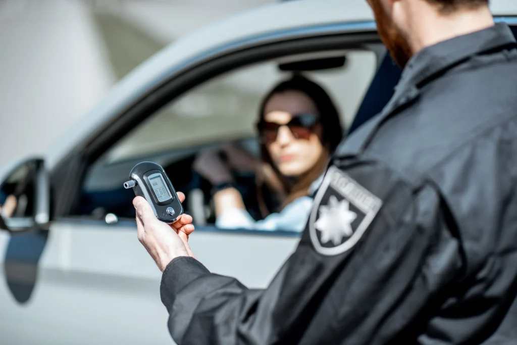 Police officer administering breathalyzer test at car window, woman wearing sunglasses in vehicle — serious sobriety check scene