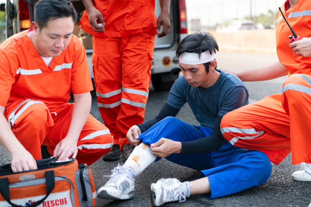 Man with a bandaged head and leg injury sits on the road as three emergency responders in orange uniforms provide first aid, conveying urgency and care.