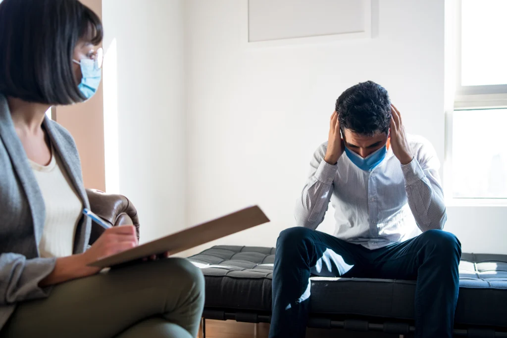 Frustrated man on couch holding head in therapy session, masked therapist taking notes in bright calm room