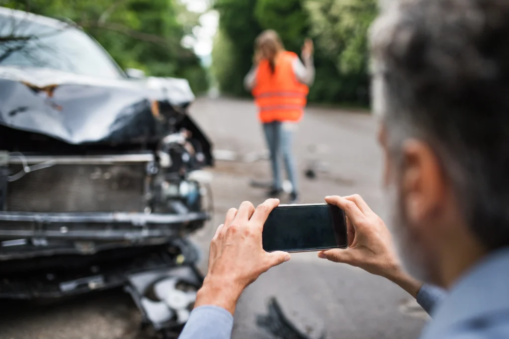 Damaged car on road with man taking photo and reflective-vest person on phone, illustrating documenting a Nevada car accident scene before filing an insurance claim.
